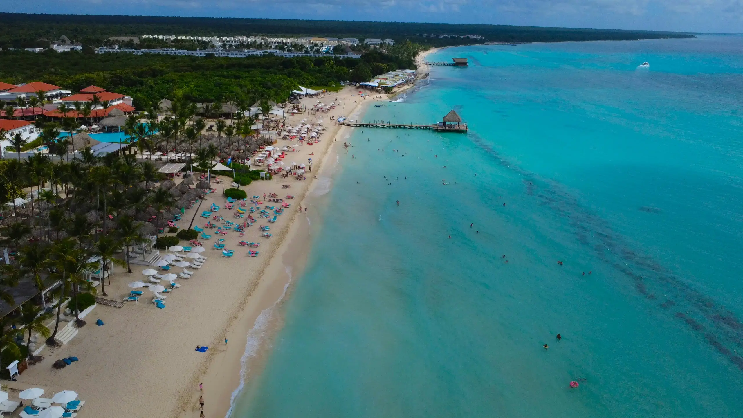 Aerial view of a resort beach in the Dominican Republic, with thatched umbrellas, a pool among palm trees and a pier over turquoise water